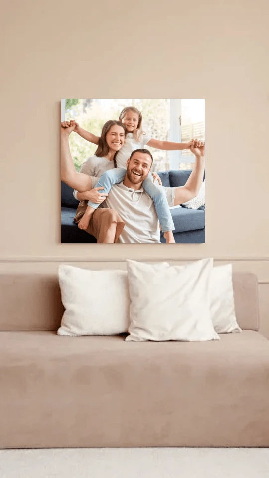 Obraz na płótnie przedstawiający uśmiechniętą rodzinę – rodziców i dziecko – siedzących na sofie w jasnym, przytulnym salonie. Dekoracja wnętrz z personalizowanym zdjęciem w tle zielonej rośliny i ceglastych poduszek.