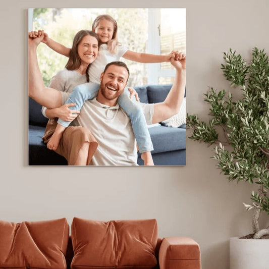 Obraz na płótnie przedstawiający uśmiechniętą rodzinę – rodziców i dziecko – siedzących na sofie w jasnym, przytulnym salonie. Dekoracja wnętrz z personalizowanym zdjęciem w tle zielonej rośliny i ceglastych poduszek.