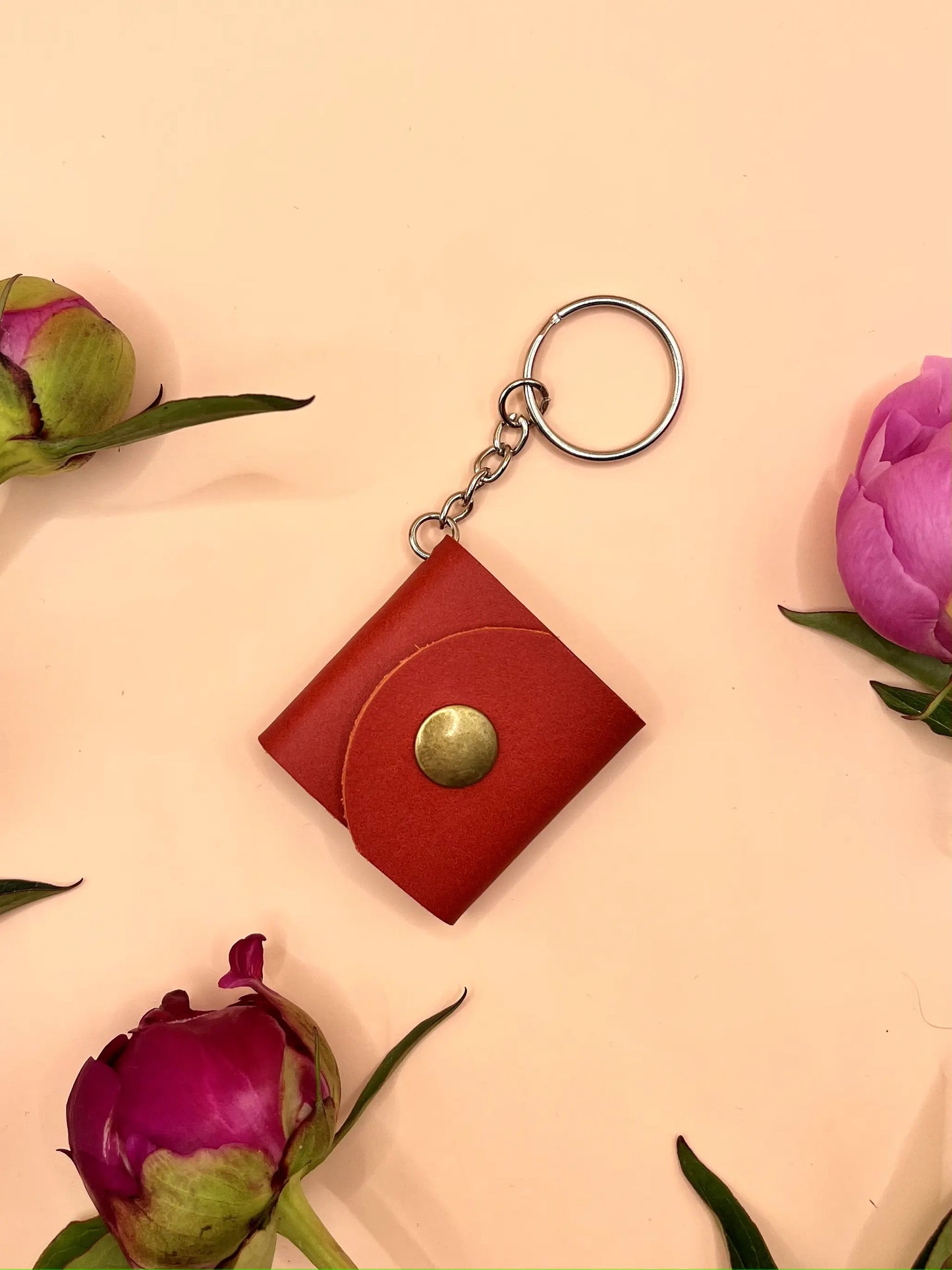 Zdjęcie przedstawia czerwony skórzany brelok z metalowym zatrzaskiem i kółkiem do przypięcia, leżący na jasnym, pastelowym tle. Wokół breloka rozmieszczone są różowe pąki piwonii, które dodają zdjęciu elegancji i delikatnego, kwiatowego charakteru.
