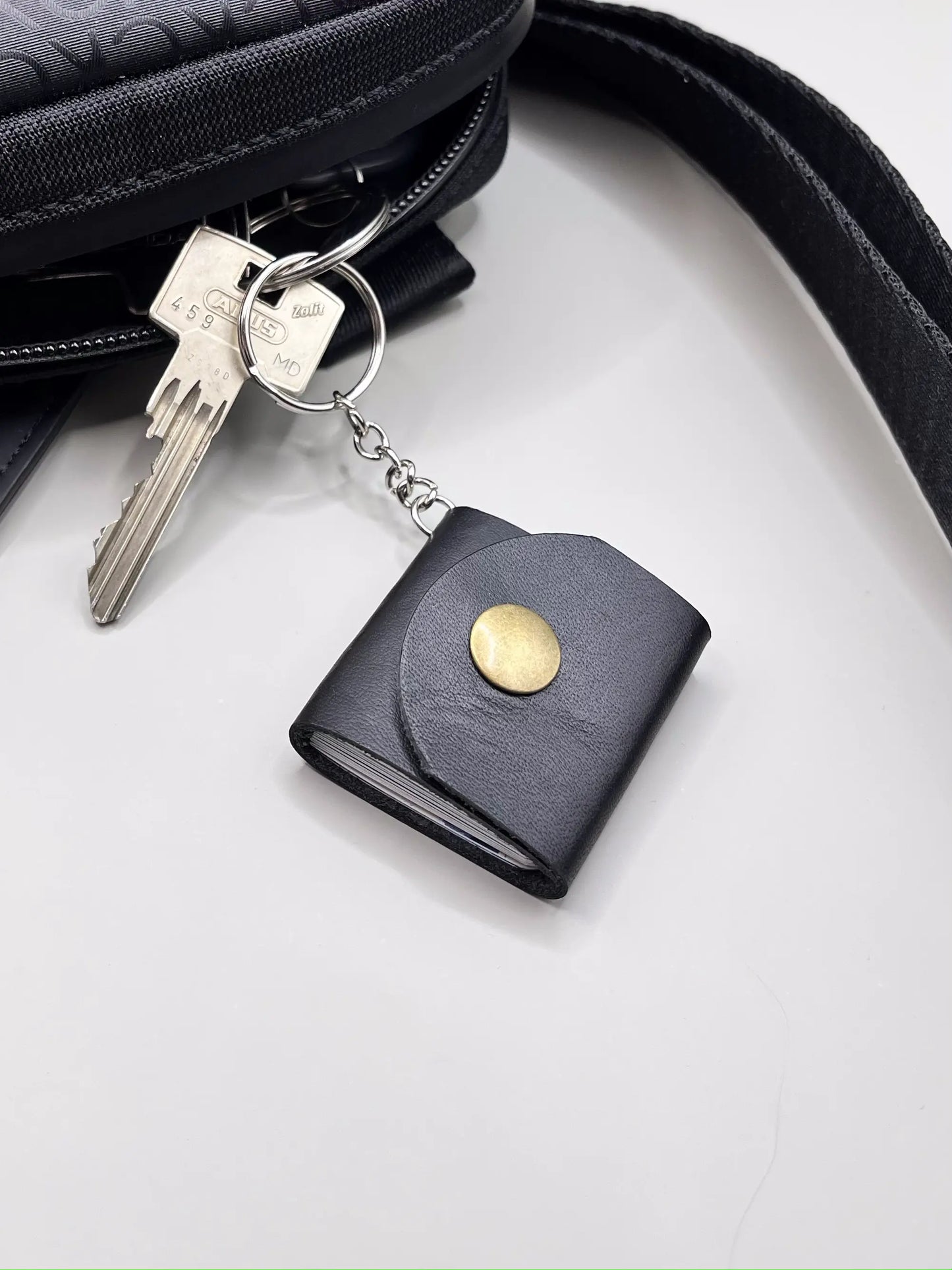 Zdjęcie przedstawia czarny skórzany brelok z metalowym zatrzaskiem, do którego przypięte są klucze. Brelok leży obok torby z czarnym paskiem, co podkreśla jego praktyczność i stylowy charakter w codziennym użytkowaniu.