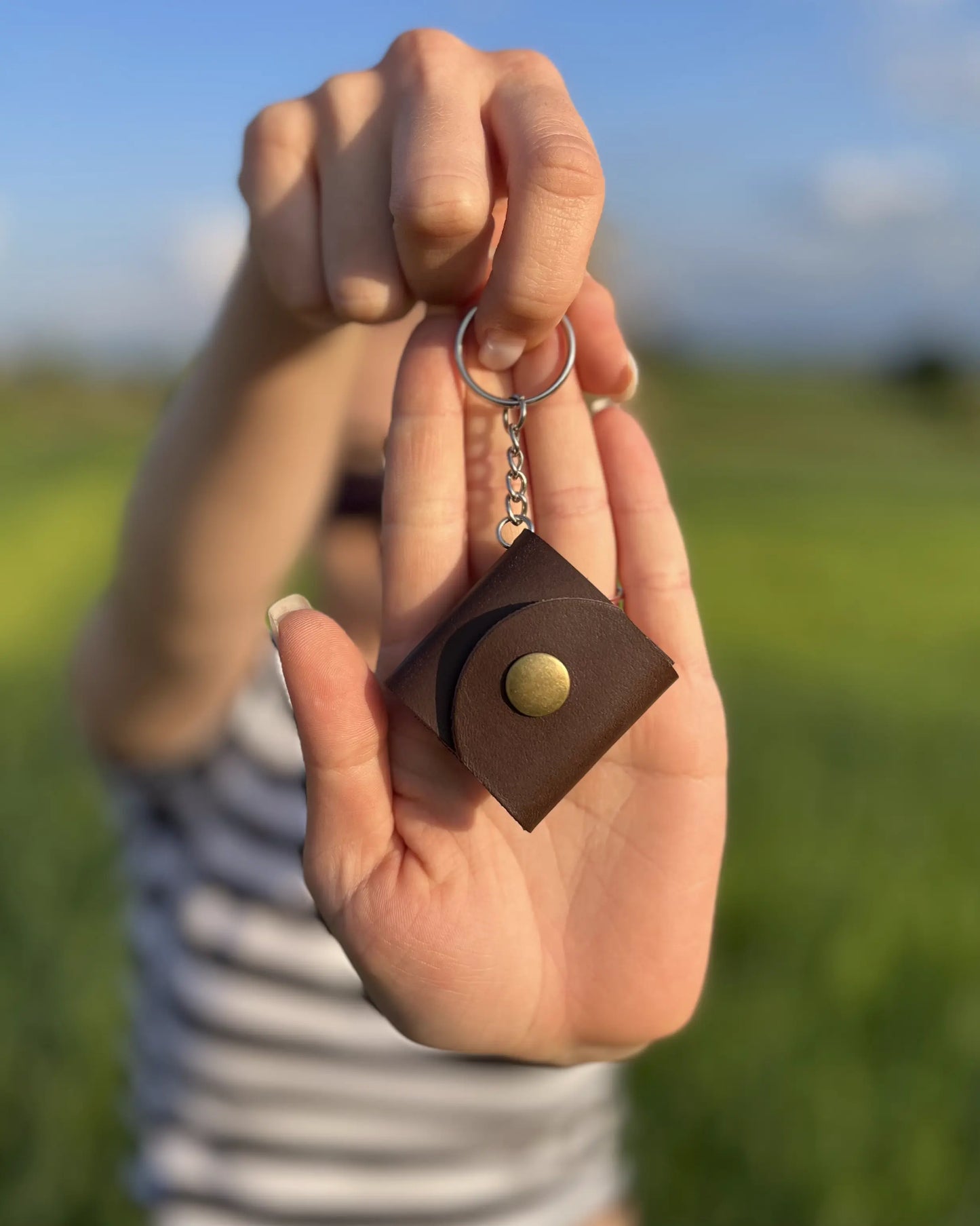 Zdjęcie przedstawia osobę trzymającą w dłoni brązowy brelok wykonany ze skóry, z metalowym zatrzaskiem i kółkiem do przypięcia. Tło stanowi rozmyty krajobraz z zielonym polem i błękitnym niebem, nadając całości naturalny i lekki charakter.