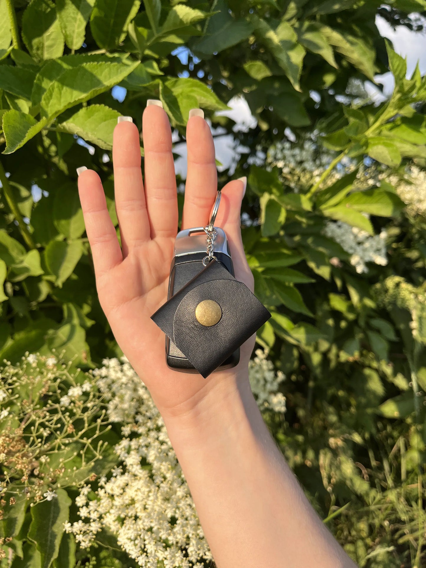 Zdjęcie przedstawia dłoń trzymającą czarny skórzany brelok z metalowym zatrzaskiem, przypięty do kluczy samochodowych. W tle widać zielone liście i białe kwiaty w naturalnym, słonecznym otoczeniu, co nadaje fotografii świeżości i organicznego charakteru."
