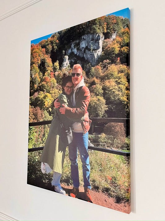 Zdjęcie przedstawia obraz na płótnie zawieszony na jasnej ścianie. Na obrazie znajduje się para przytulająca się na tle jesiennego krajobrazu z kolorowymi drzewami i skałami. Obraz charakteryzuje się żywymi barwami i wysoką jakością wydruku.