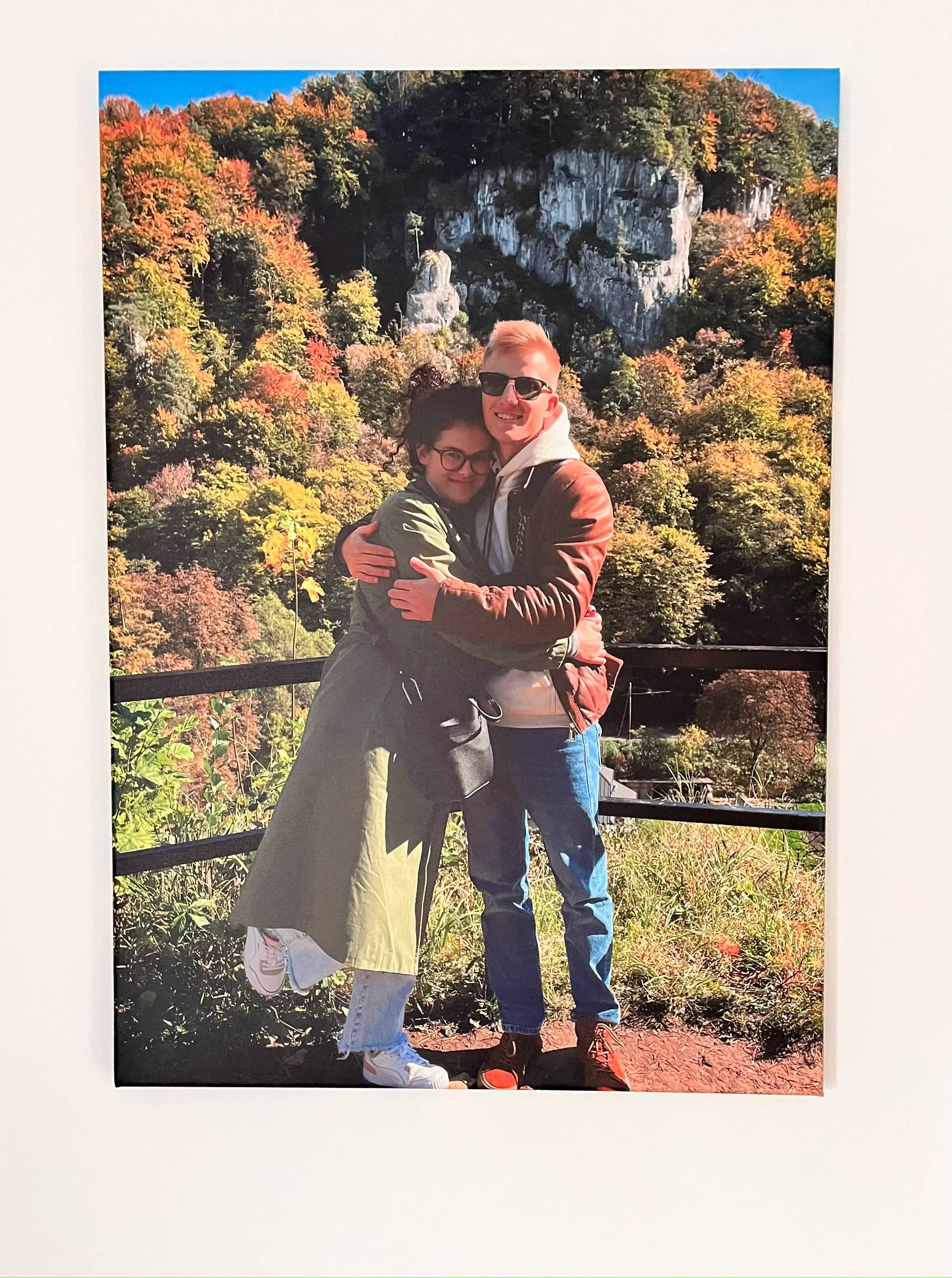 Zdjęcie przedstawia obraz na płótnie zawieszony na jasnej ścianie. Na obrazie znajduje się para przytulająca się na tle jesiennego krajobrazu z kolorowymi drzewami i skałami. Obraz charakteryzuje się żywymi barwami i wysoką jakością wydruku.