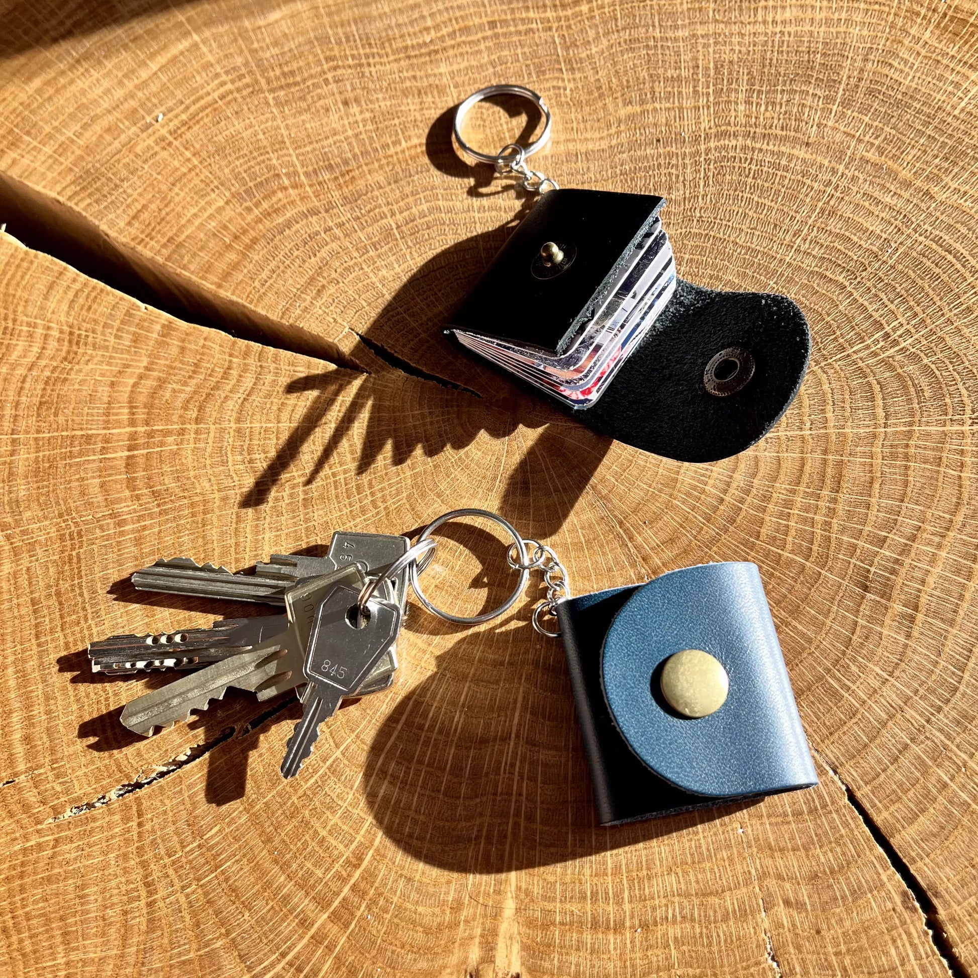 Zdjęcie przedstawia dwa breloki leżące na drewnianej powierzchni z widocznymi słojami drewna. Jeden z breloków, w czarnym kolorze, jest otwarty i ukazuje przechowywane w środku zdjęcia. Drugi brelok, w kolorze granatowym, jest zamknięty i posiada metalowy zatrzask. Do breloka przypięte są klucze, co pokazuje jego funkcjonalność jako breloka do kluczy