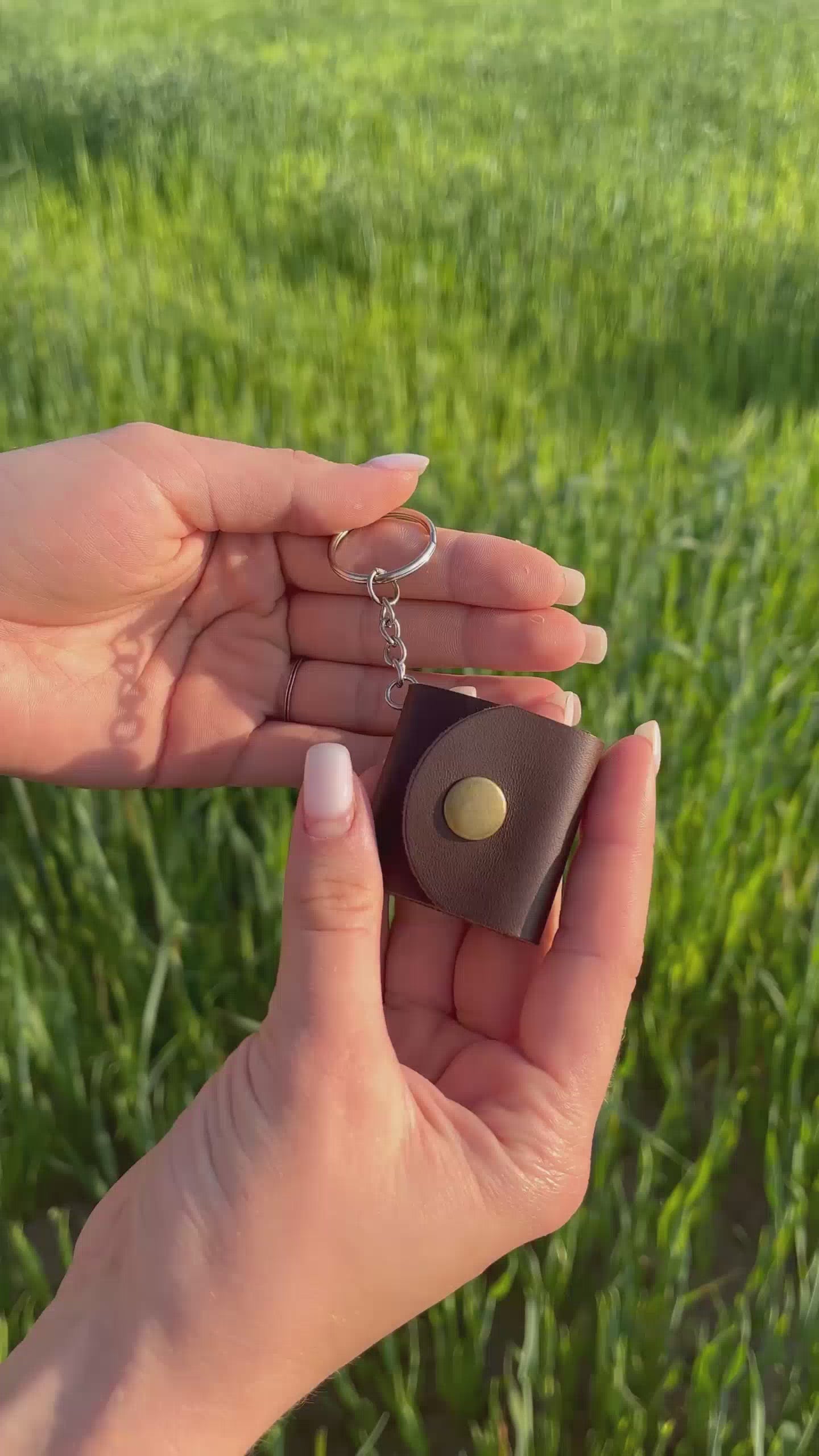 Brązowy skórzany brelok ze zdjęciami — mini album na zdjęcia personalizowany, idealny na prezent. Filmik ukazuje elegancki brelok otwierany na zatrzask, z miejscem na kilka fotografii, prezentowany na tle zielonej łąki.