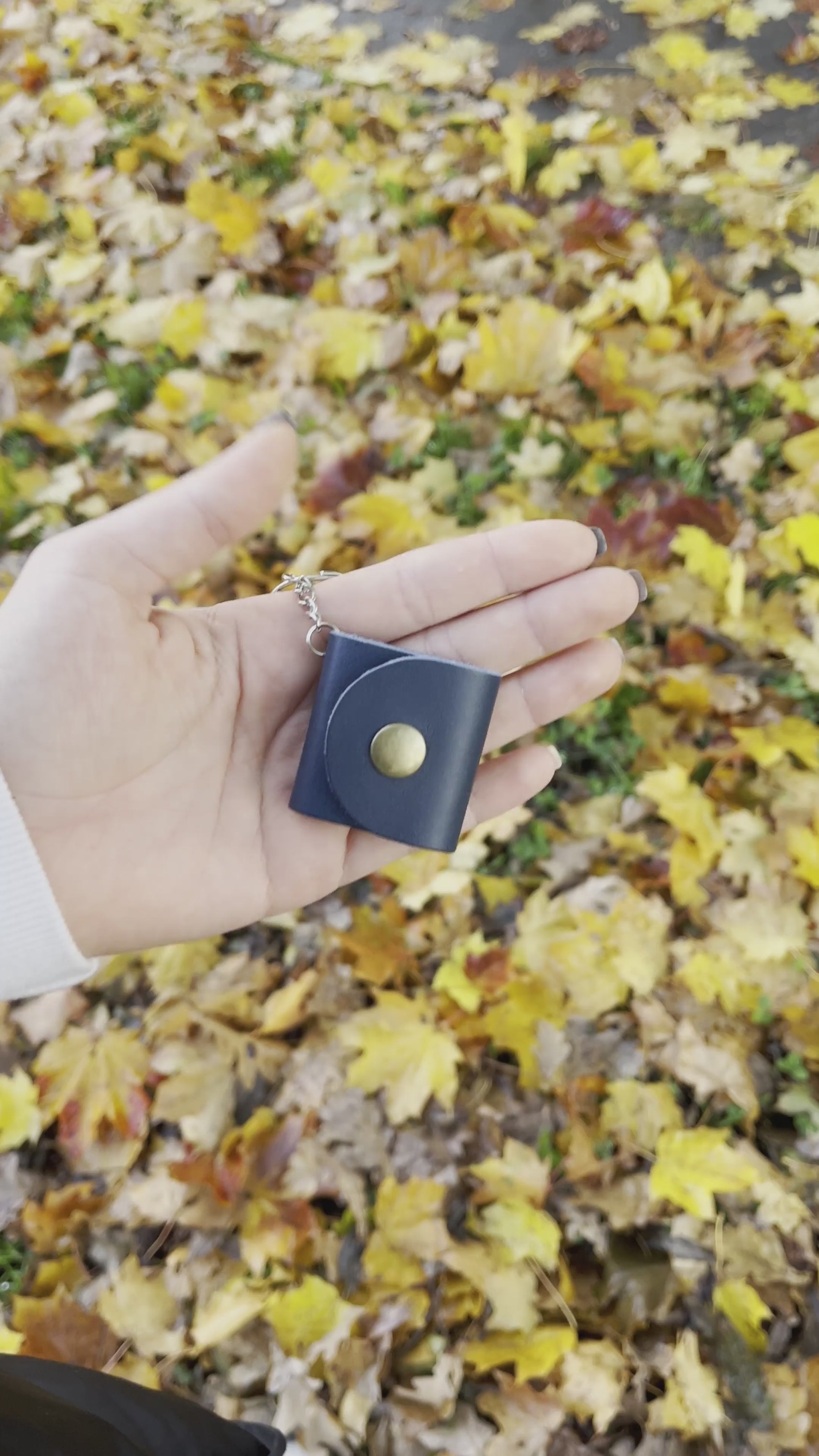 Nagranie prezentuje granatowy brelok wykonany ze skóry, który delikatnie się otwiera, ukazując w środku zdjęcia. W tle widać jesienną scenerię – na ziemi leżą żółte liście, które dodają ciepłego, naturalnego klimatu nagraniu.