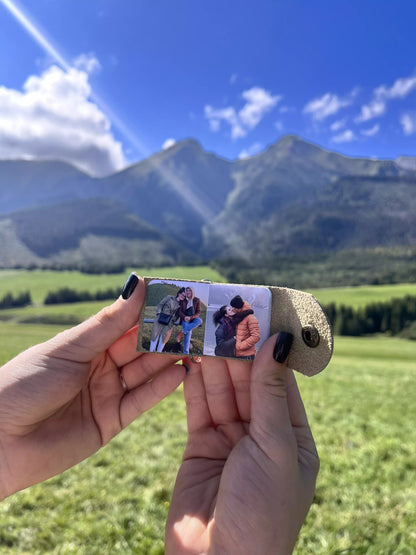 Zdjęcie przedstawia ręce trzymające otwarty brelok w jasnym kolorze, ukazujący dwa personalizowane zdjęcia w środku. W tle widoczny jest malowniczy krajobraz z zieloną łąką, majestatycznymi górami i błękitnym niebem z białymi chmurami, co nadaje fotografii wyjątkowego, naturalnego uroku.