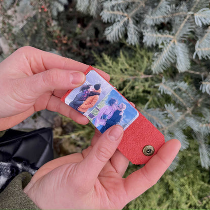 Zdjęcie przedstawia ręce trzymające czerwony skórzany brelok, który jest otwarty, ukazując dwa personalizowane zdjęcia w środku. W tle widoczne są zielone gałązki świerku, co nadaje fotografii zimowego, naturalnego klimatu.